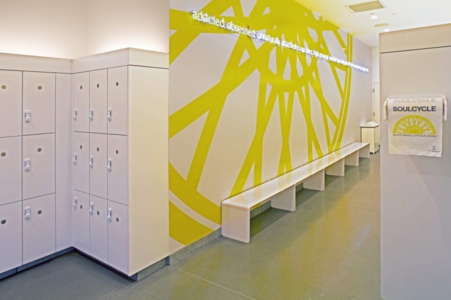 White laminate lockers, a wall featuring yellow SoulCycle logo with neon text on top and a long wall bench.